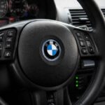 Czarna kierownica BMW z przyciskami sterującymi i deską rozdzielczą w tle
