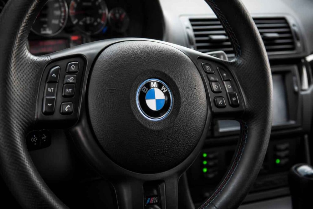 Czarna kierownica BMW z przyciskami sterującymi i deską rozdzielczą w tle