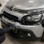 Mechanik w rękawicach montuje przedni zderzak srebrnego samochodu Citroën w warsztacie