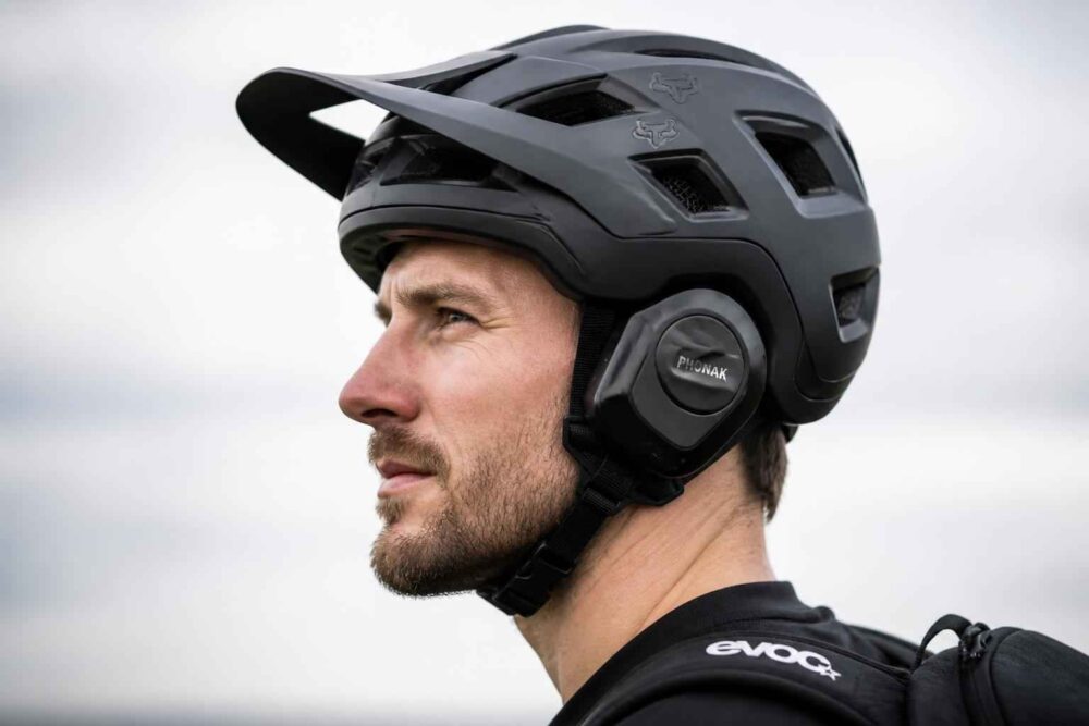 Mężczyzna w czarnym kasku rowerowym z daszkiem i ochroną uszu przy plecaku evoke