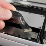 Dłoń podnosi elastyczną taśmę połączeniową wewnątrz rozebranego urządzenia elektronicznego